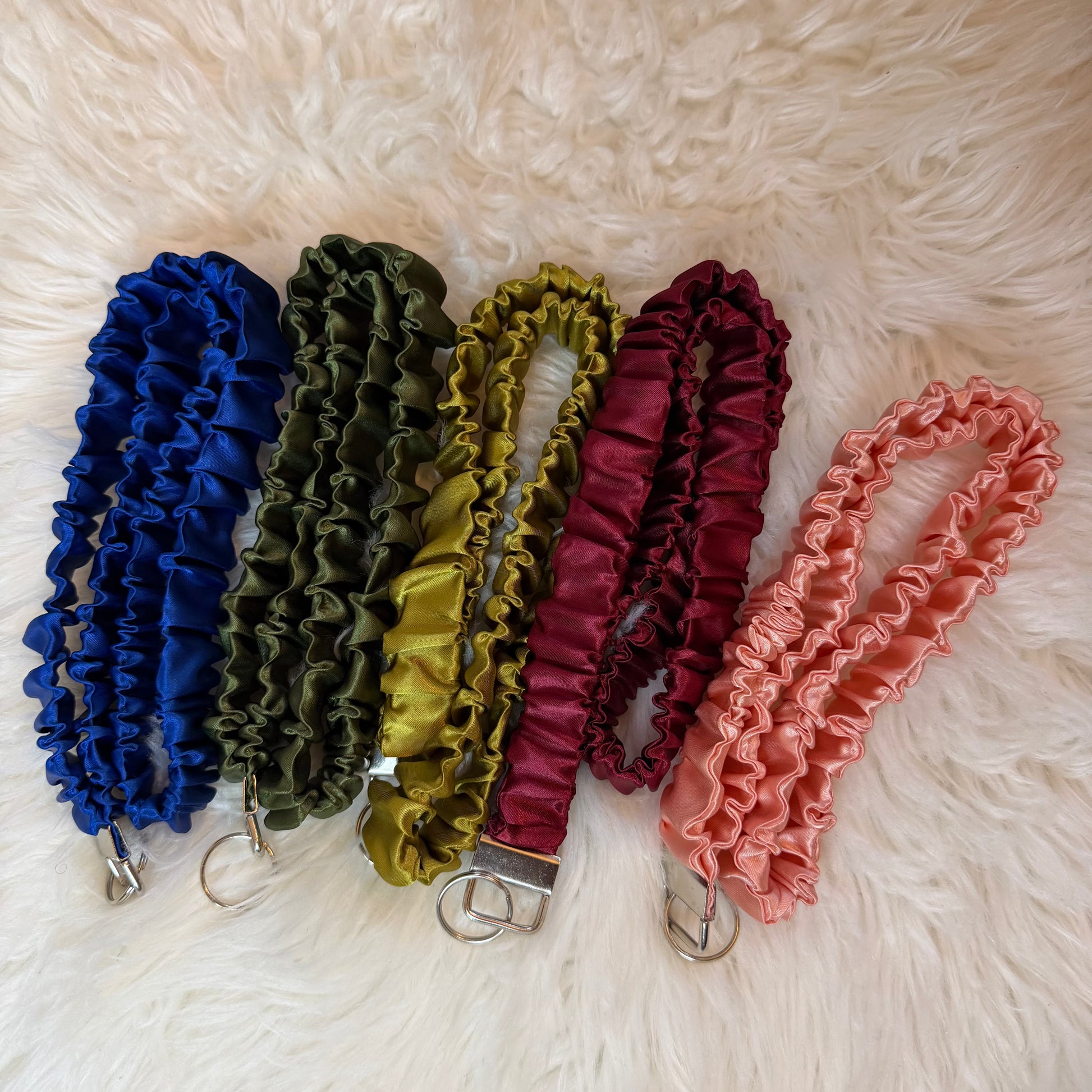 Five satin wristlets in blue, olive green, mustard yellow, burgundy, and dusty pink, arranged on a white faux fur background. Each wristlet has a silver metal clasp and ring.